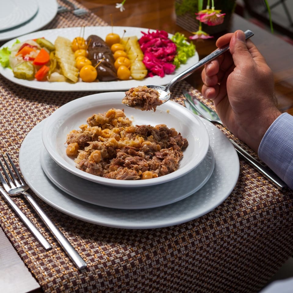 A person spoons a Peruvian dish of shredded meat and chickpeas from a white bowl, with pickled vegetables in the background.